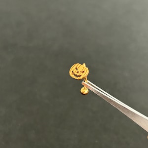 Puede incluir: Pendiente de calabaza de color dorado con una cara de calabaza tallada. El pendiente est&aacute; sujeto por unas pinzas.