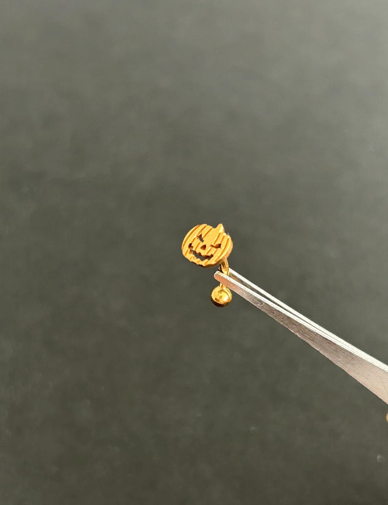 Puede incluir: Pendiente de calabaza de color dorado con una cara de calabaza tallada. El pendiente est&aacute; sujeto por unas pinzas.