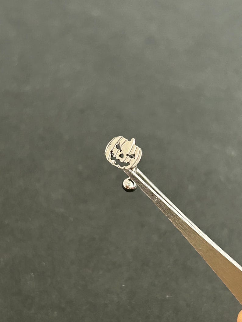 Puede incluir: Un charm de calabaza plateado con una cara tallada, unido a una bola plateada en una cadena plateada.
