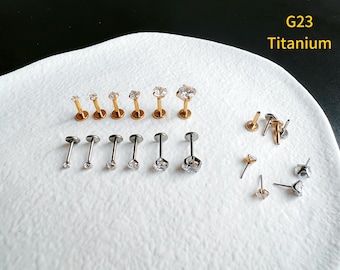 Pode incluir: Close-up de uma variedade de joias corporais de titânio com pedras de zircônia cúbica transparentes. As joias estão dispostas sobre uma superfície branca. G23 Titânio.