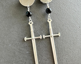 May include: A pair of silver earrings with a disc top, a black bead, and a sword pendant. The earrings have a medieval theme and a polished finish. The sword pendants are approximately 7.6 cm long.