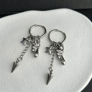 May include: A pair of silver hoop earrings with dangling charms. Each earring features a star, small beads, a teardrop shape, and a chain ending in a cone-shaped spike. The earrings are displayed on a white surface.