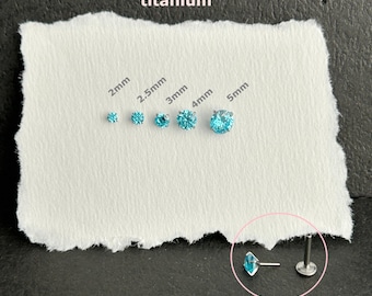 Pode incluir: Um conjunto de cinco brincos de titânio com pedras redondas azul-brilhante, organizados por tamanho de 2mm a 5mm. O texto titanium está impresso acima. Um brinco é mostrado de lado, revelando um pino de base plana e a cravação.