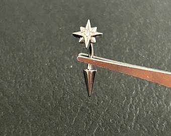 Può includere: Un gioiello per il corpo a barra curva color argento, caratterizzato da una parte superiore a forma di stella con una piccola pietra chiara incastonata e un'estremità inferiore a forma di cono appuntito. Il metallo ha una finitura lucida ed è tenuto da una pinzetta.