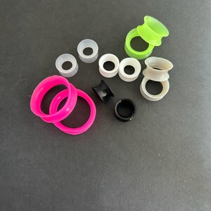 May include: A collection of colorful ear gauges in various sizes and styles. The gauges include bright pink, neon green, black, white, and gray silicone tunnels and plugs. The assortment is displayed against a dark background.