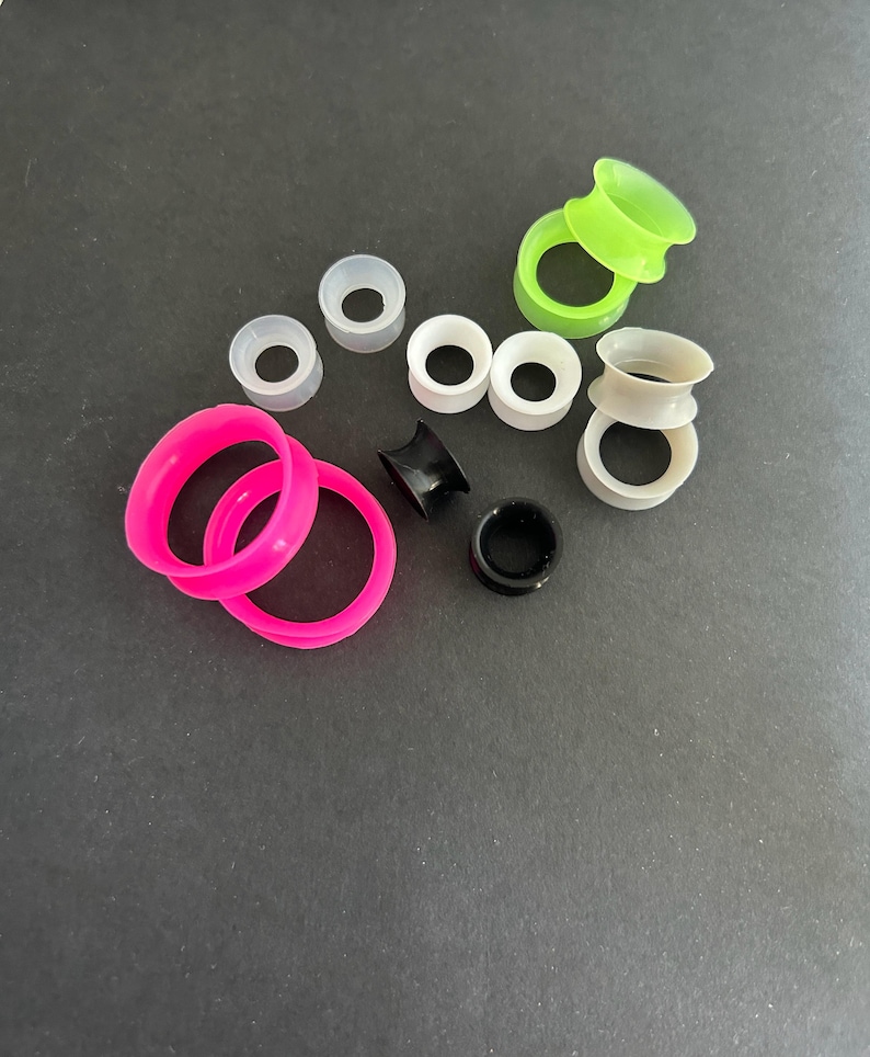 May include: A collection of colorful ear gauges in various sizes and styles. The gauges include bright pink, neon green, black, white, and gray silicone tunnels and plugs. The assortment is displayed against a dark background.