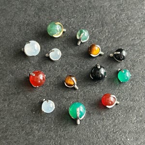 May include: Assortment of colorful gemstone beads with silver settings. The beads are in various shapes and sizes, including round, oval, and teardrop. The beads are arranged in a circle on a dark gray surface.