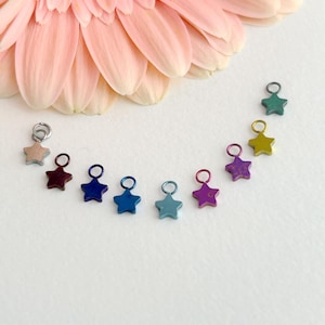 May include: A collection of colorful star-shaped charms in various hues, including blue, purple, and green, arranged in a semi-circle. The charms are small, metallic, and have a loop at the top. A large, light pink flower is in the background.