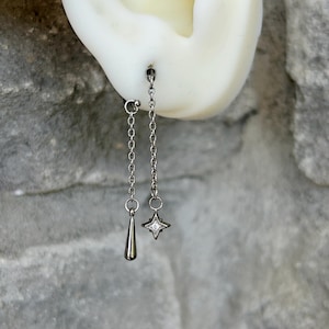 Op de afbeelding: Zilveren oorhangers. Eén oorbel heeft een druppelvormige bedel, de andere een ster met een centrale heldere steen. Beide hangen aan fijne kettingen. De oorbellen worden tegen een neutrale achtergrond getoond.