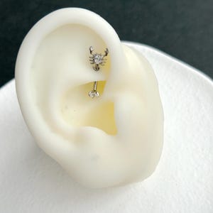 Pode incluir: Uma joia de piercing de barra curva em tom prateado com um topo em forma de escorpião adornado com uma pedra transparente e uma esfera a condizer com uma pedra transparente na base, exibida num modelo de orelha branco para mostrar a colocação.
