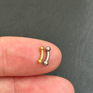 Op de afbeelding: Twee gebogen barbell piercings. Eén is goudkleurig en de andere zilverkleurig. Elk heeft aan beide uiteinden een kleine bal. De sieraden worden op een vingertop getoond tegen een donkere achtergrond.