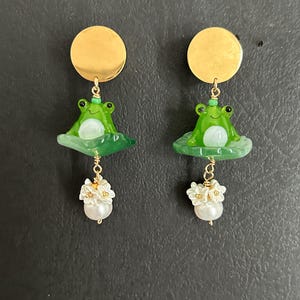 May include: A pair of gold frog earrings with white pearl and flower accents. The frogs are green with black eyes and sit on a green leaf.