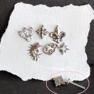 May include: A collection of seven silver-toned metal body jewelry pieces, including a marquise-cut crystal cluster, a four-pointed star, a fleur-de-lis, a bird with a red stone eye, a cloud shape, a star, and a flat-back stud with a post.