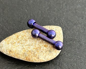 May include: Two purple barbell body jewelry pieces with rounded ends. The jewelry is on a small, gray rock.