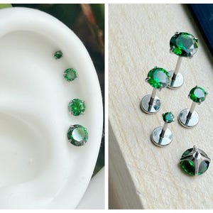 Op de afbeelding: Vijf lichaamssieraden met groene edelstenen en zilveren pinnen. De edelstenen zijn van verschillende grootte en zijn rond gezet. De sieraden worden getoond op een wit oppervlak en op het oor van een persoon.