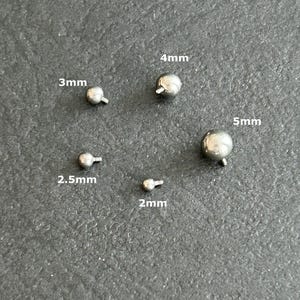 Op de afbeelding: Een verzameling van vijf kleine, zilverkleurige, kogelvormige sieraden met schroefpalen. Maten zijn gelabeld als 2 mm, 2,5 mm, 3 mm, 4 mm en 5 mm. De stukken worden weergegeven op een donkergrijze ondergrond.