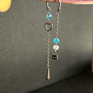 Könnte beinhalten: Ein silberfarbener Ohrring mit einer gebogenen Creolenbasis und zwei hängenden Ketten. Eine Kette trägt ein kleines Herz-Element und einen tropfenförmigen Anhänger, die andere zwei leuchtend blaue Perlen, eine klare Perle und einen schwarzen Katzenanhänger.