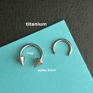 May include: Two silver-colored titanium body jewelry pieces. One is a horseshoe-shaped ring with pointed spikes, labeled "spike 5mm" and "14g". The other is a smaller, curved ring. The word "titanium" is also visible.
