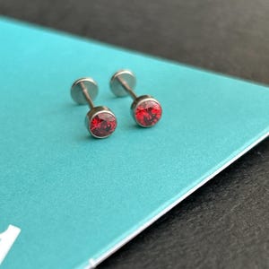 Pode incluir: Dois pequenos brincos de pressão prateados com pedras preciosas redondas vermelhas brilhantes. Os brincos são exibidos em uma superfície azul-turquesa, com um fundo escuro. Os brincos têm um design simples e minimalista.