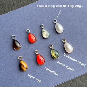 May include: A collection of eight teardrop-shaped gemstone charms with metal jump rings. The stones include tiger eye, carnelian, moss agate, and imitation pearl. Text on the image states: This O ring will fit 18g 20g, tiger eye, carnelian, moss agate, imitation pearl.