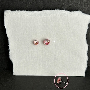May include: Two small, round, pink and red gemstone stud earrings on a white surface. The earrings are set in a silver metal. The image is taken from a high angle, with a dark background.