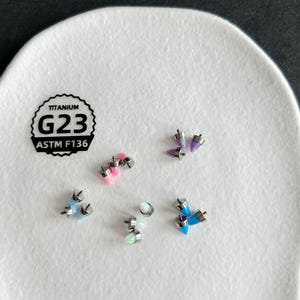 May include: A collection of small, cone-shaped body jewelry attachments in various colors including pink, purple, blue, and white. Each piece features a metallic base and is labeled with a black stamp reading TITANIUM G23 ASTM F136.