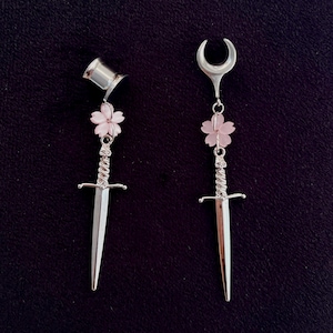 Pode incluir: Um par de brincos prateados com um design de espada. Cada brinco apresenta uma pequena flor rosa e uma pega decorativa. Um tem um topo cilíndrico, o outro uma forma de meia-lua.