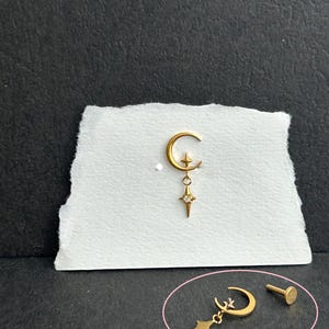 Può includere: Un gioiello per il corpo color oro con un ciondolo a forma di mezzaluna e una piccola stella a quattro punte fissata al centro. Una stella a quattro punte con una pietra chiara è appesa alla luna. È incluso anche un perno a base piatta.