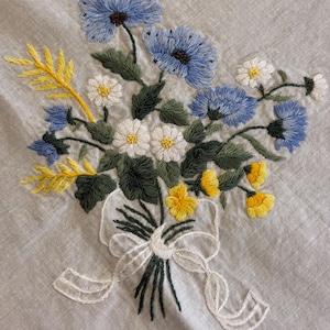 May include: Embroidered floral bouquet featuring blue, white, and yellow flowers with green leaves. The bouquet is tied with a white ribbon. The embroidery is on a light gray fabric.