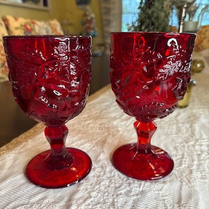 May include: Two ruby red goblets with intricate floral and leaf patterns. The goblets have a round base, a stem, and a detailed bowl. The glassware is made of a transparent material, allowing light to pass through. The goblets are placed on a white, textured surface.