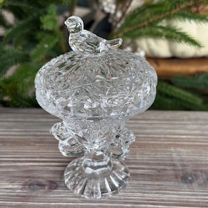 May include: A clear pressed glass compote dish featuring a faceted diamond pattern, a pedestal base, and a lid topped with a small bird figurine. The glass bowl has a scalloped rim and decorative glass accents around the exterior of the stem.