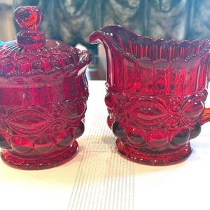 May include: A ruby red glass sugar bowl with a matching lid and a creamer pitcher. Both pieces feature a pressed glass pattern with circular indentations and decorative scrollwork, finished with a scalloped rim and a sturdy handle on the creamer.