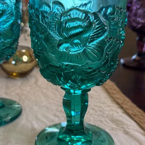 May include: A teal-colored glass goblet with a detailed rose pattern. The stem and base are also teal, with a floral design on the base. The glass has a vintage aesthetic, with a clear, textured appearance.