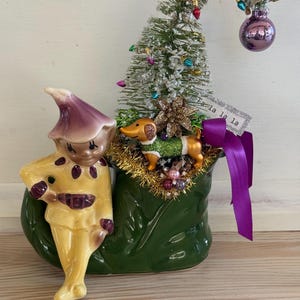 May include: A ceramic elf figurine in a yellow outfit with a purple hat sits on a green boot-shaped planter. The planter holds a small flocked Christmas tree with colorful lights, a purple ornament, a dachshund figurine, and a sign reading Fa la la la.