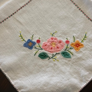 May include: A white linen tablecloth with embroidered floral details. The embroidery features a central arrangement of pink, blue, and orange flowers with green leaves and red berries. The border is trimmed with a green and red stitched pattern.