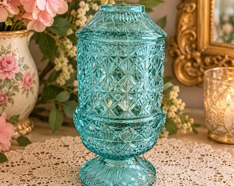 May include: A teal blue glass jar with a lid, featuring a textured diamond and starburst pattern across its cylindrical body, a flared base, and a domed top. The glass is translucent and reflects light across its faceted surface.