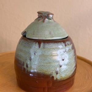 May include: A handmade ceramic jar with a lid. The jar is a rounded shape with a brown base and a green and brown upper section. The lid is green with a small insect-shaped handle. The jar is sitting on a wooden tray.