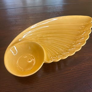 May include: A yellow ceramic serving dish with a unique, wing-like design. The dish features a round bowl on one end and a long, curved section with a scalloped edge. The surface has a glossy finish and a textured, ridged pattern.