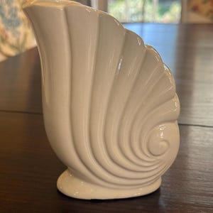 May include: A cream-colored, seashell-shaped ceramic pitcher. The pitcher has a fluted design on the body and a rounded base. The spout is on the left side. The pitcher is a decorative item.