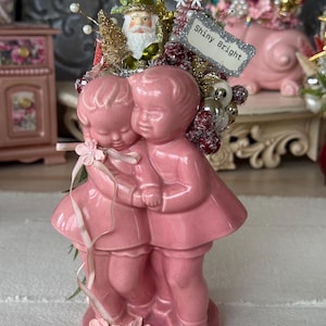 May include: A pink ceramic figurine of two children embracing, adorned with a decorative arrangement featuring a Santa ornament, a silver sign reading Shiny Bright, glittery gold accents, white berries, and a small pink ribbon bow.