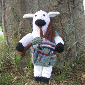 May include: A white knitted sheep toy with black eyes and a brown nose, wearing a green, blue, and red plaid kilt. The sheep is holding a brown stick in its mouth.