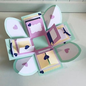 May include: An open, multi-layered paper craft box in pastel colors. The box features light green, white, pink, and yellow elements, with heart-shaped accents. It has multiple compartments and decorative details.