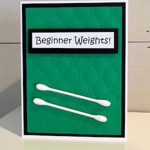 May include: A handmade greeting card with a green textured background and a black border. The card features the text "Beginner Weights!" in a white rectangle and two white cotton swabs.