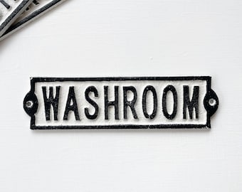 May include: A white cast iron sign with black lettering that reads "WASHROOM". The sign has two small holes on either side for hanging.