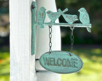 May include: A teal blue metal welcome sign with four birds perched on a branch. The sign is hanging from a white wooden post and says "WELCOME" in a rustic font.