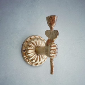 May include: Gold metal wall hook in the shape of a hand holding a rose. The hook is mounted on a circular base with a decorative design.