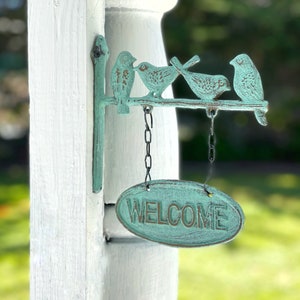 May include: A teal blue metal welcome sign with four birds perched on a branch. The sign is hanging from a white wooden post and says "WELCOME" in a rustic font.