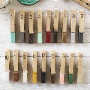 May include: Eighteen wooden clothespins painted in various colors, numbered 1 through 18. The clothespins are arranged in two rows of nine on a white wooden surface.