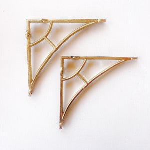 May include: Two gold metal shelf brackets with a decorative, geometric design. The brackets are curved and have a traditional style.
