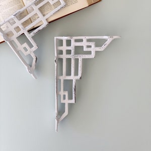 May include: Two white metal shelf brackets with a geometric design. The brackets are made of cast iron and have a distressed finish.